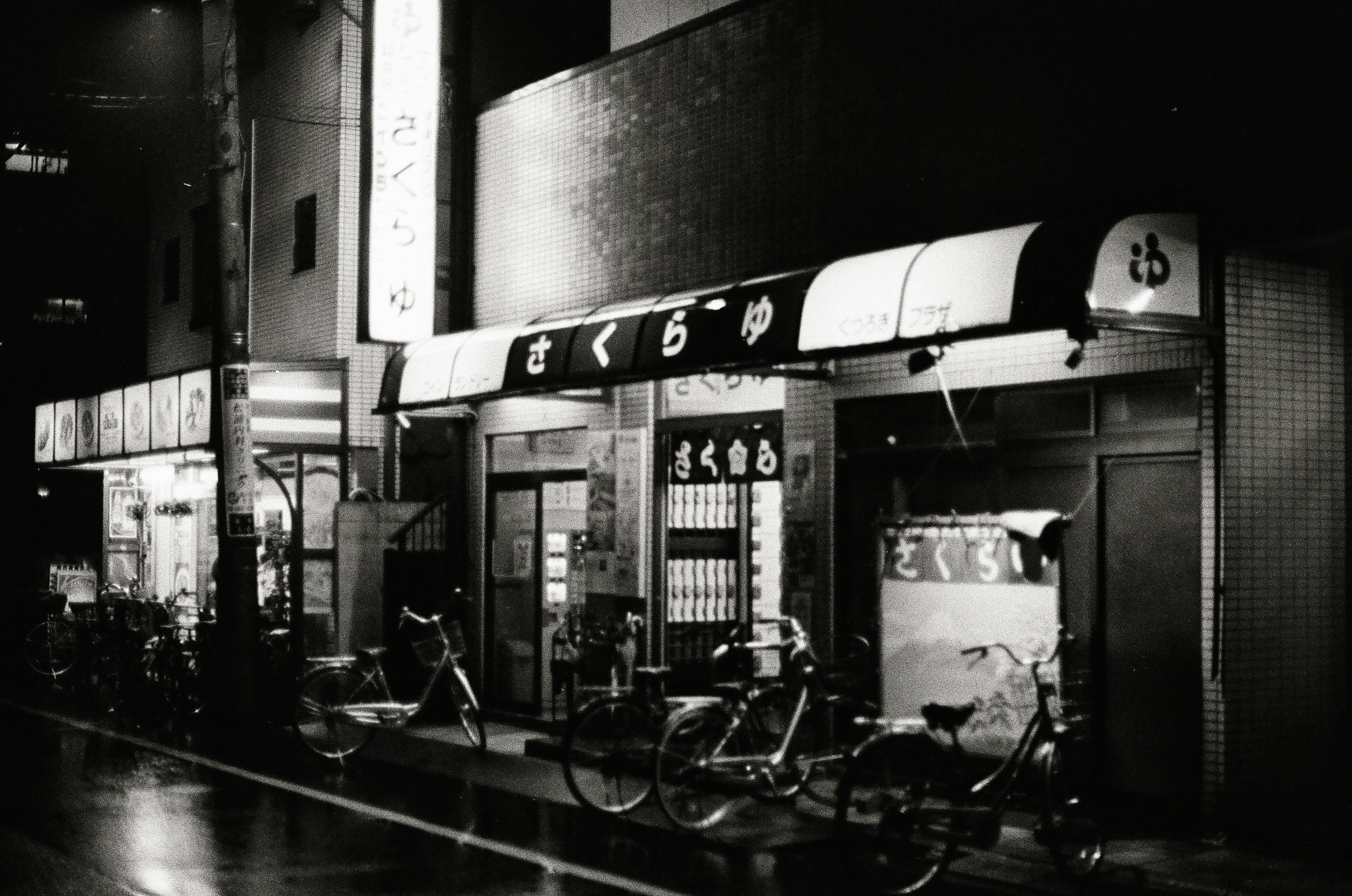 Bicycles are parked outside a brightly lit Japanese bath house. A sign with the name of the venue is above the door. A laundry room is lit up on the left of the entrance.