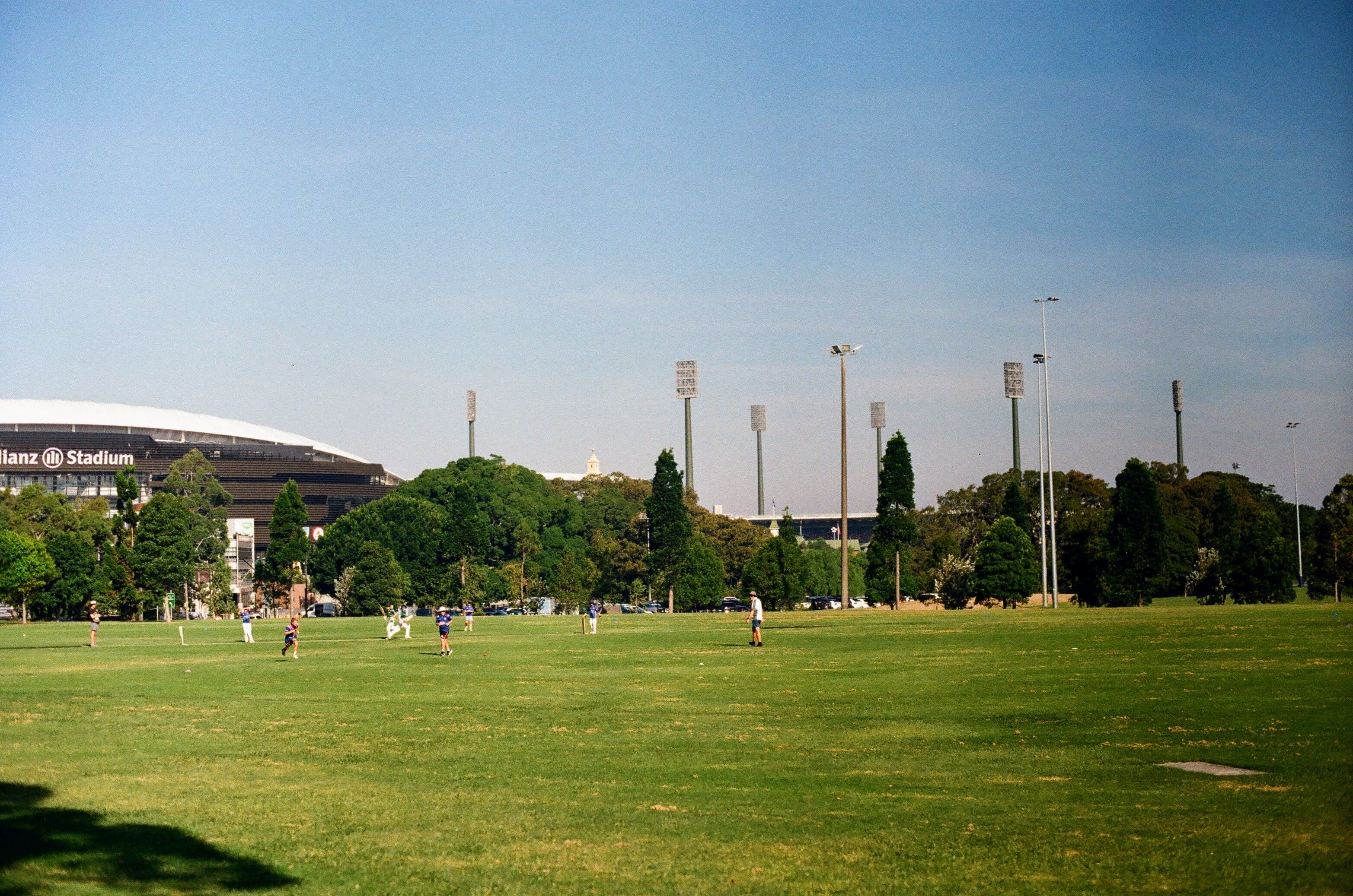 Kids playing cricket in a large open field, with blue skies above. Two large stadiums dominate the background of the shot.