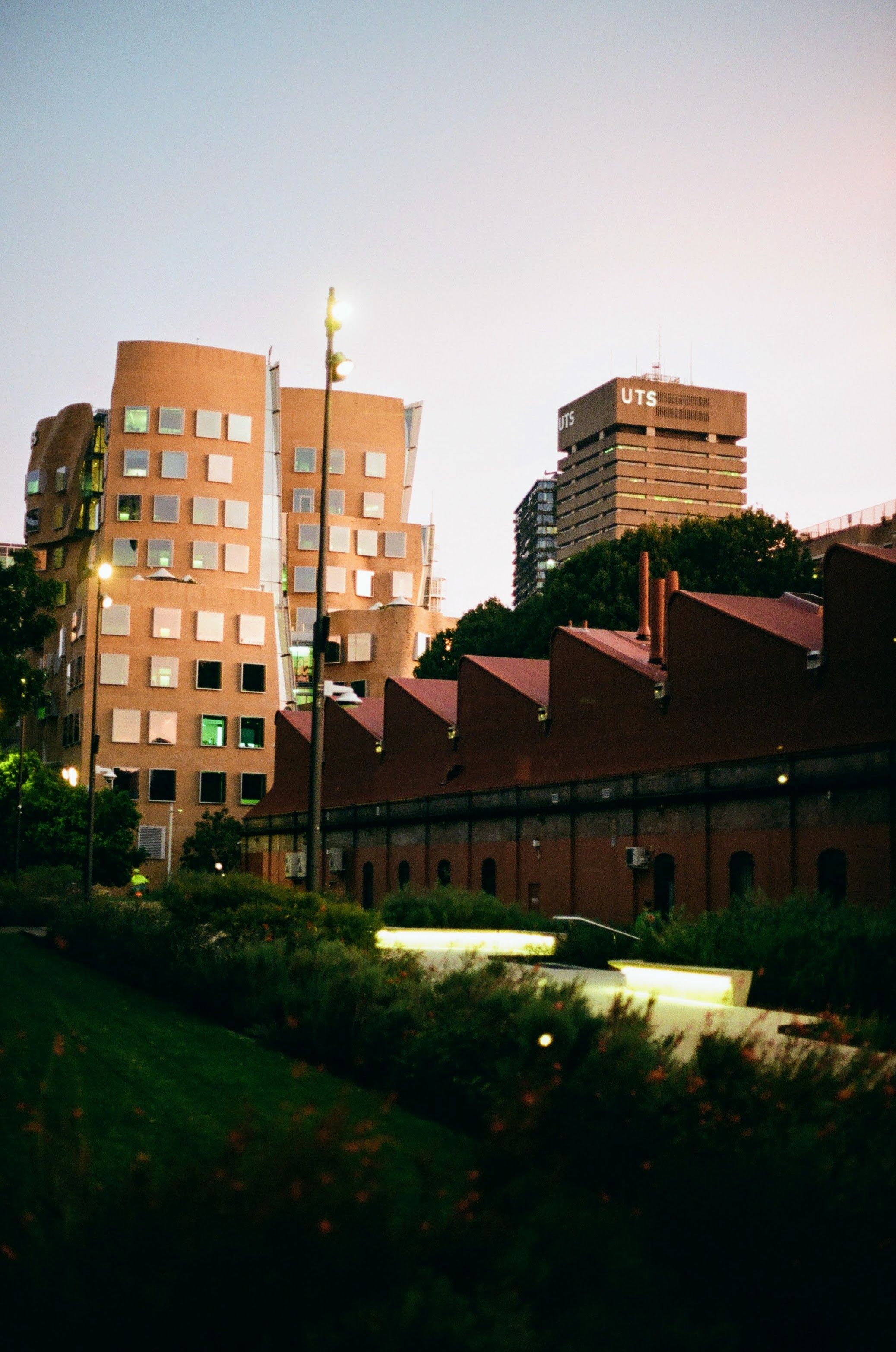 An old industrial building is on the right of the shot, alongside a garden walkway. In the background is a newly designed university building on the left, with an older building to the right of it.